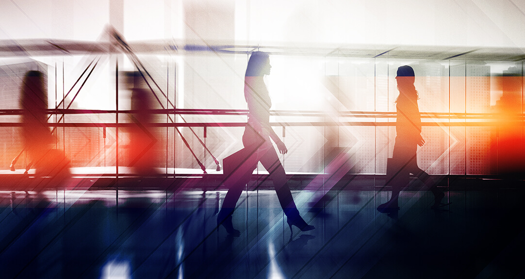 Businesspeople walk the halls of an office building as sunlight filters through the windows, creating a warm, glowing effect.