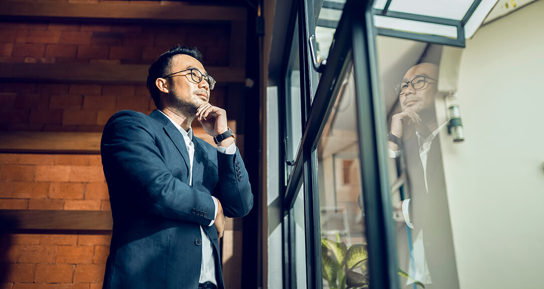 An Asian businessman thoughtfully looks through a large window, his reflection evident in the glass.