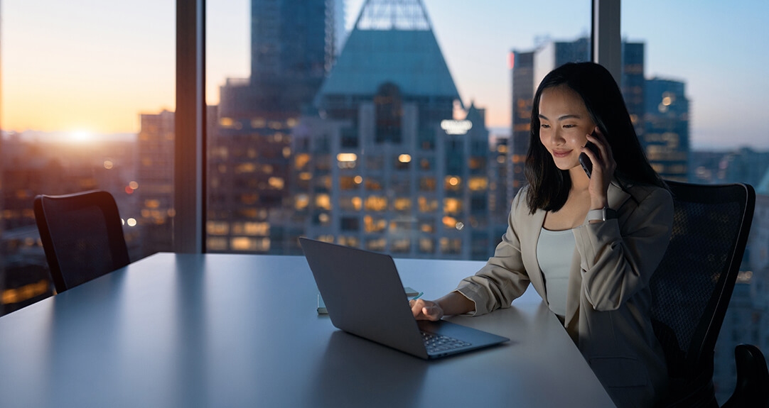 A woman in a business suit works at a laptop while she uses her smartphone.