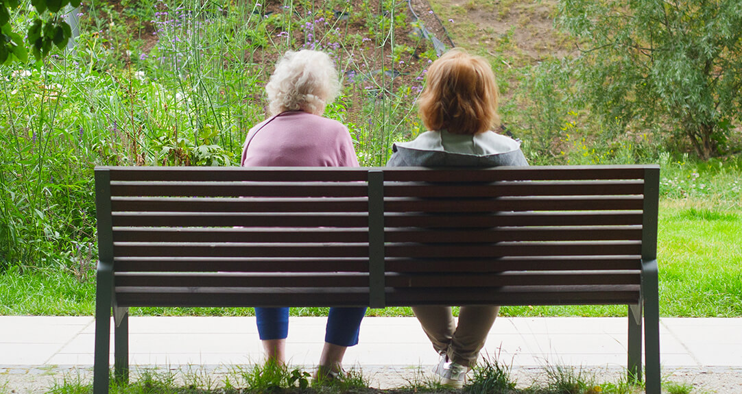An elderly woman and a younger woman sot on a park bench with their backs to the viewer.