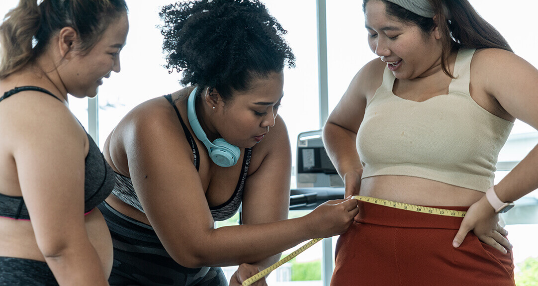 Two overweight women in workout clothes check the waist measurements of a third.