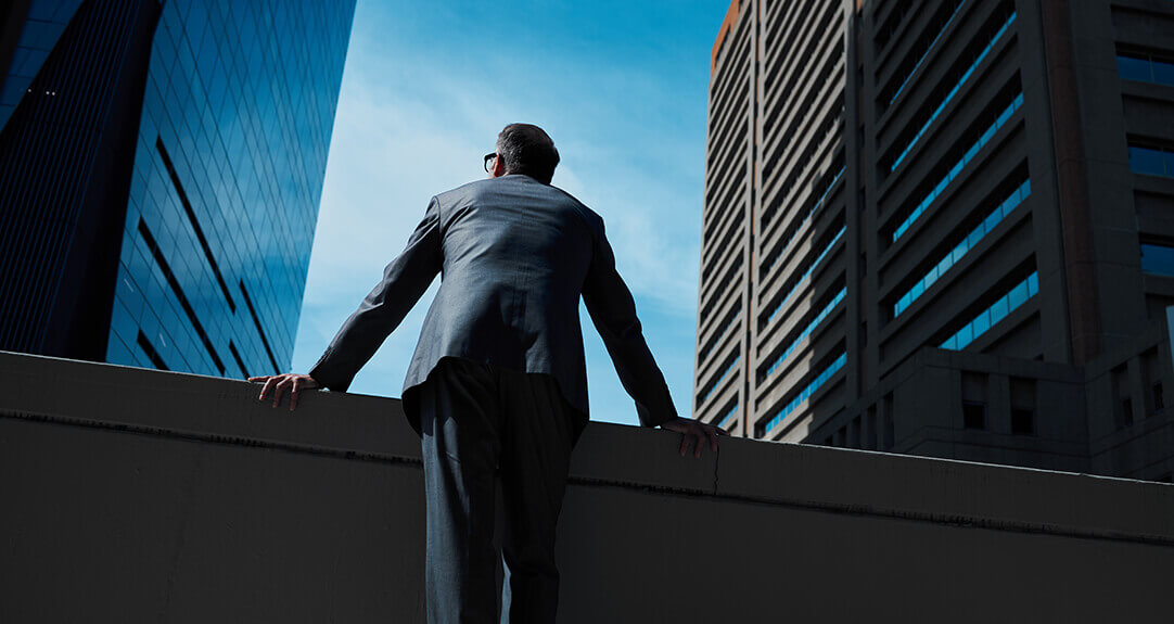 An older man in a suit faces away from the viewer and looks at a vista of office buildings.