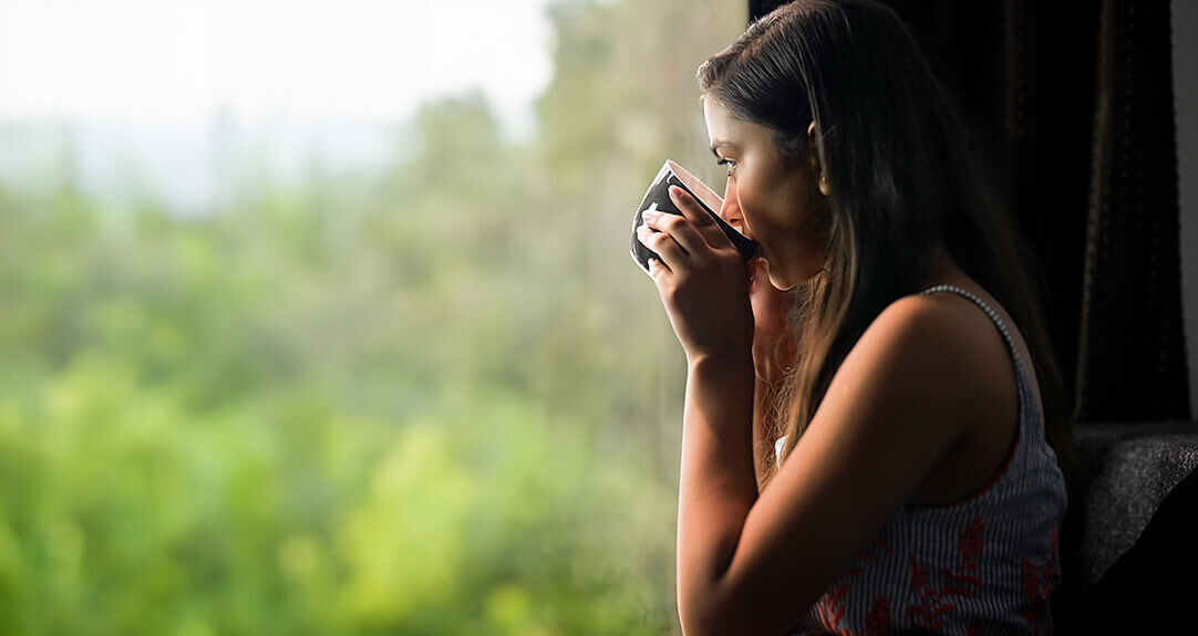 A woman sits in a dark room sipping tea and looking outside at a green landscape.