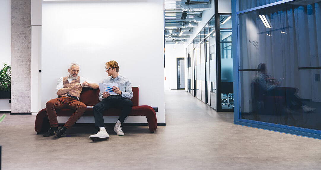 Two men seated on an office sofa discuss their work.