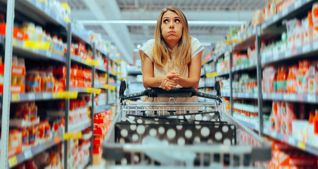 A woman looks bewildered as she pushes a shopping cart down a grocery store aisle.